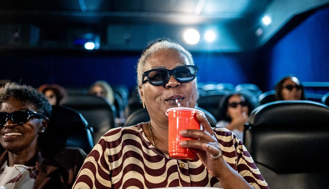 Mature woman drinking soda and wearing 3-D glasses in a dark movie theater with lights from the projector in the background